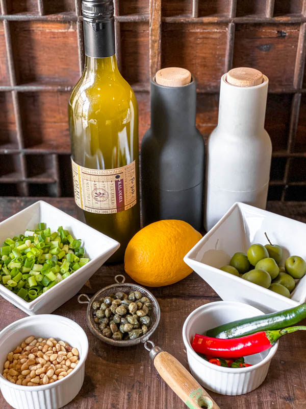 A photo showing all the ingredients required to make the Salmon Tartare dressing: a bottle of olive oil, salt and pepper shakers, sicilian olives, chillies, a lemon, a small strainer with salted capers, toasted pinenuts, chopped spring onions.