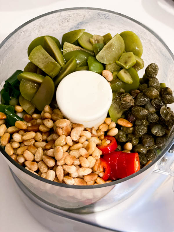 A small food processor bowl filled with toasted pinenuts, chopped red chillies, salted capers, chopped sicilian olives and spring onions, ready to be blitzed.