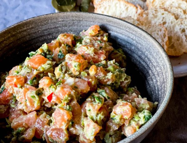 A bowl of Salmon Tartare on a grey tablecloth with thinly sliced bread rounds behind it on a plate.