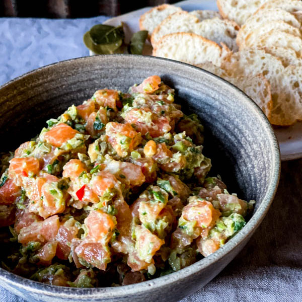 A bowl of Salmon Tartare on a grey tablecloth with thinly sliced bread rounds behind it on a plate.