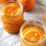 Two jars of confit orange slices in syrup with whole oranges in the background.