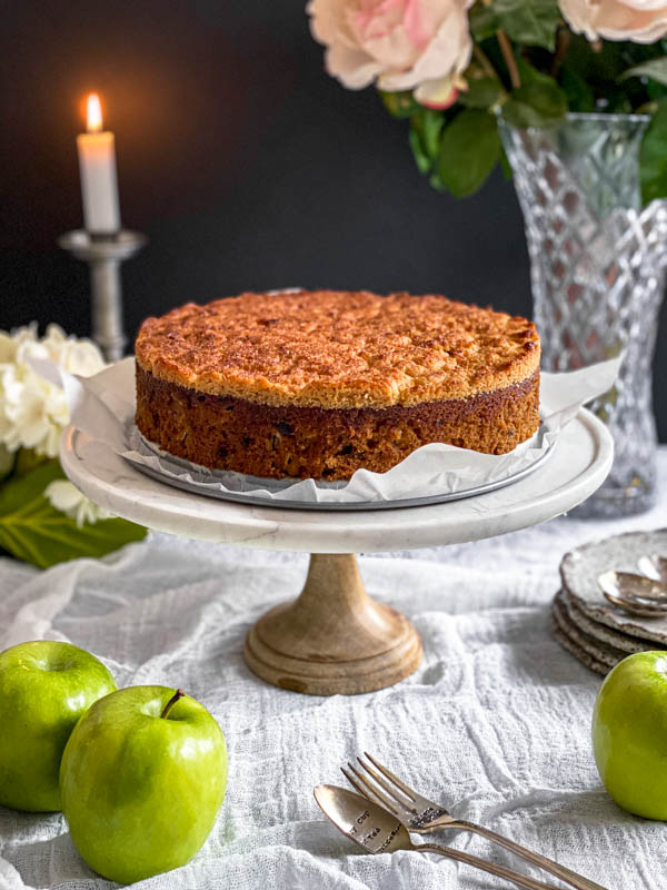 Here the Lumberjack Cake is on a marble cake stand with a wooden base. It is on a table with a candle burning in the background and a vase of flowers. There are also green apples in the foreground with cutlery and side plates off to the side.