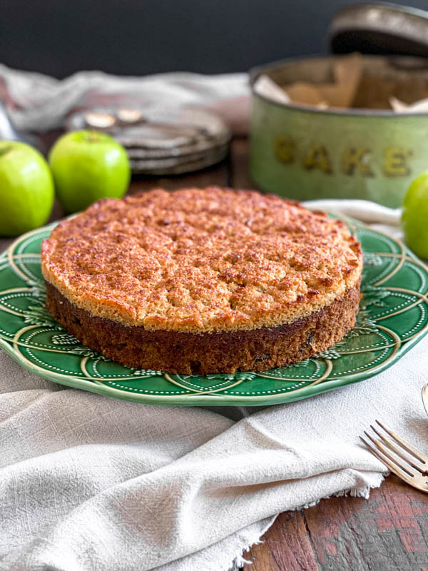 A close up of the Lumberjack Cake on a decorative green plate. It is set on a wooden table with a neutral linen tablecloth bunched under it. There are green apples in the background along with an old green cake tin.