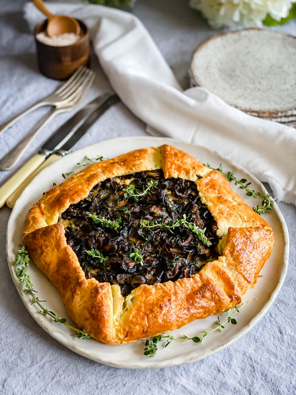 The Mushroom, Ricotta and Gruyere Galette is on a large plate surrounded by thyme sprigs. In the background are knives, forks and serving plates as well as a salt pinch-pot.