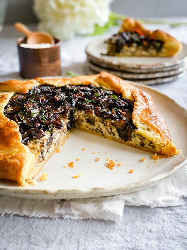 The galette has been sliced and you can now see the layers inside the galette. The cheese layer is visible, as is the mushroom layer on top. Behind the tart is a stack of serving plates with a slice of galette on the top plate. There is also a little wooden pinch-pot of sea salt beside them.