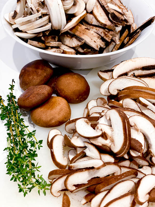 The mushrooms are all sliced. Some are in a bowl and some on the bench. There are also a few whole shitake mushrooms and some thyme sprigs in the photo.