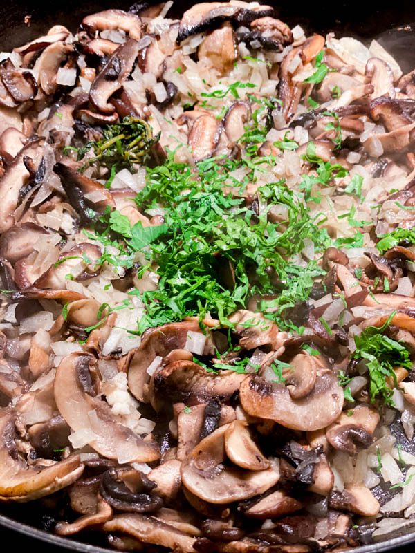 The mushrooms are being sautéed in a large frying pan, along with onions, garlic and parsley has just been added.