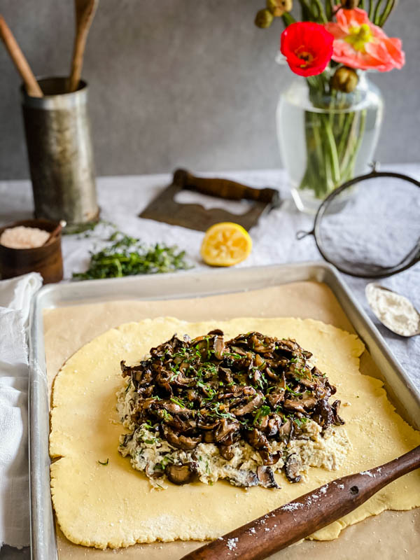 The mushrooms are now on top of the cheese mixture and ready for the pastry sides to be folded over. It is on a baking paper lined oven tray on a table with kitchen utensils around it.