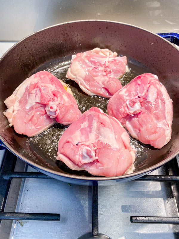 The raw bone-in chicken thighs are being browned skin-side down in a sauté pan.