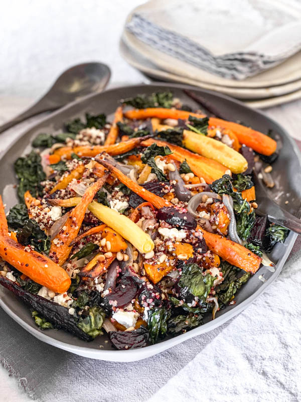 A platter of Roasted Vegetable Salad with Quinoa and Feta, on a table with a serving spoon to the side and plates and napkins in the background.