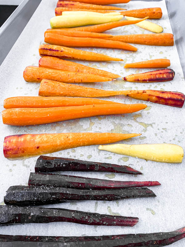 Baby heirloom carrots that have been oiled and salted are spread out on an oven tray.