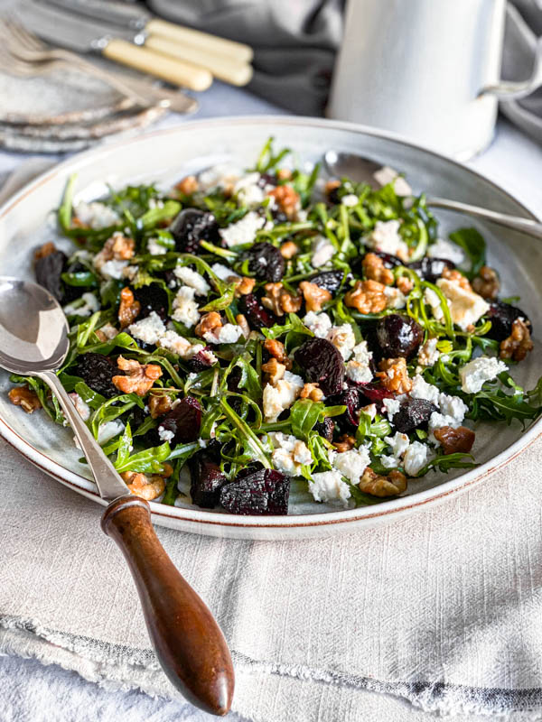 The salad is on a table with salad servers on the platter. In the background are plates, cutlery and a white jug/