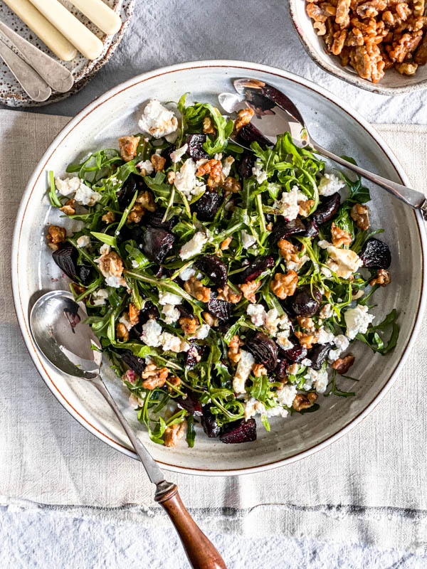 Looking down onto the Roast Beetroot, Goats Cheese and Walnut Salad. There are salad servers on the platter and at the back are a bowl of candied walnuts and cutlery.