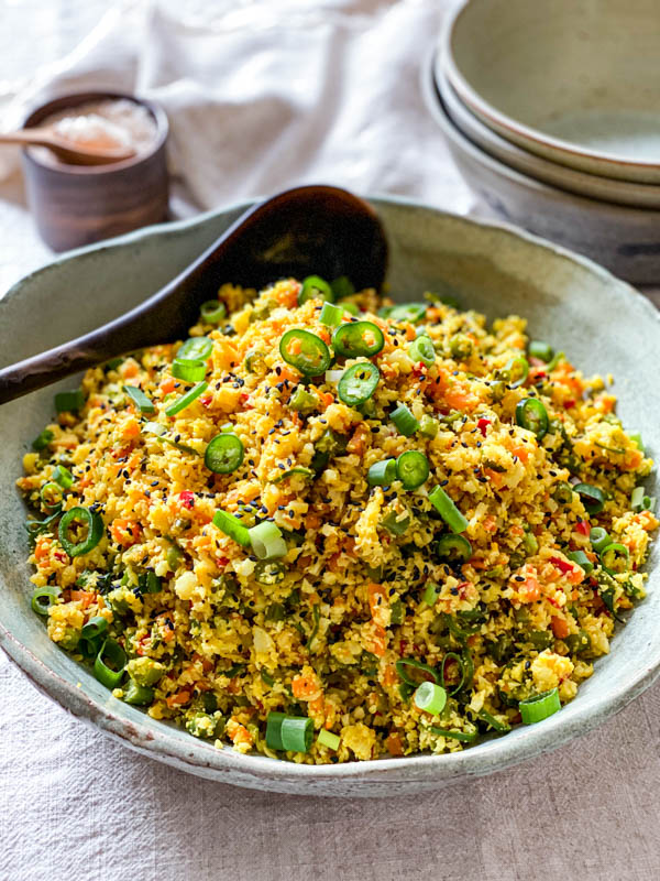 The Cauliflower Fried Rice with Turmeric is in a green ceramic bowl on a table with a serving spoon. In the background are 3 stacked bowls and a pinch bowl of salt.