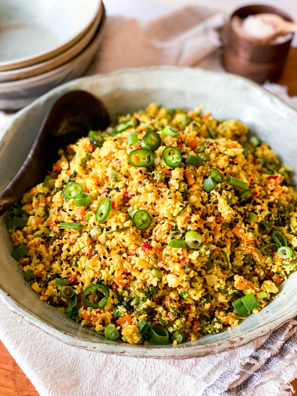 The Cauliflower Fried Rice with Turmeric is in a serving bowl on a folded napkin with bowls and salt in the background.