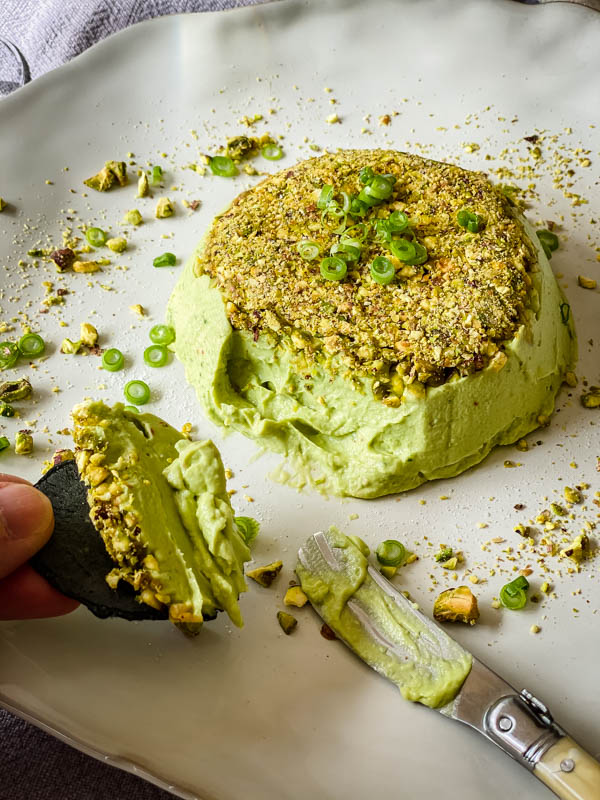 A cracker with some of the pate is being held with the Avocado Pistachio Pate in the background.