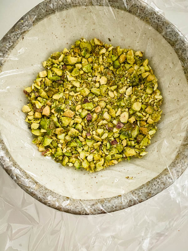 The chopped pistachio nuts are in a small bowl that has been lined with cling wrap.