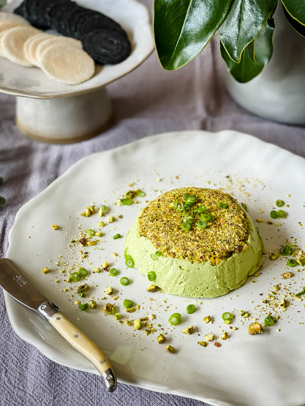 The platter with the Avocado Pistachio Pate is in on a table with crackers on a plate behind it.