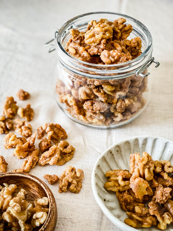A jar of Candied Walnuts in the back with some scattered in front and two small bowls of nuts in the foreground.