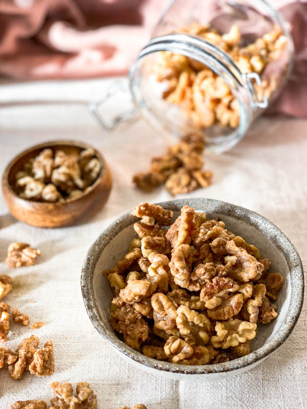 A bowl of Candied Walnuts with a jar in the background that is tipped over with some of the nuts spilling out onto the tablecloth.