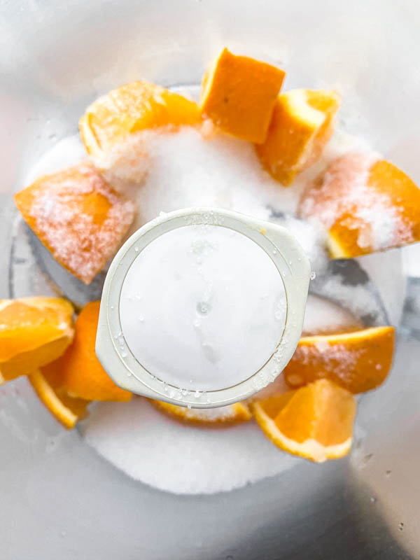 The chopped orange chunks are in the bowl of a food processor with the sugar ready to be blended.