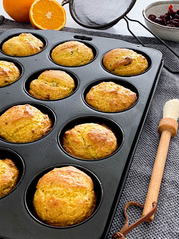 A tray of muffins cooling. The muffin tray is on a tea towel on a kitchen bench with a pastry brush beside it and a sieve, an orange half and a bowl of cranberries in the background.