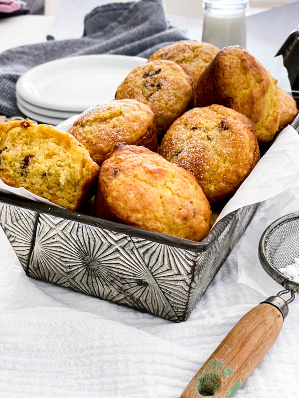 A loaf tin lined with baking paper is full of freshly baked muffins. There are plates and a glass of milk in the background and a small sieve with icing sugar in it, in the foreground.