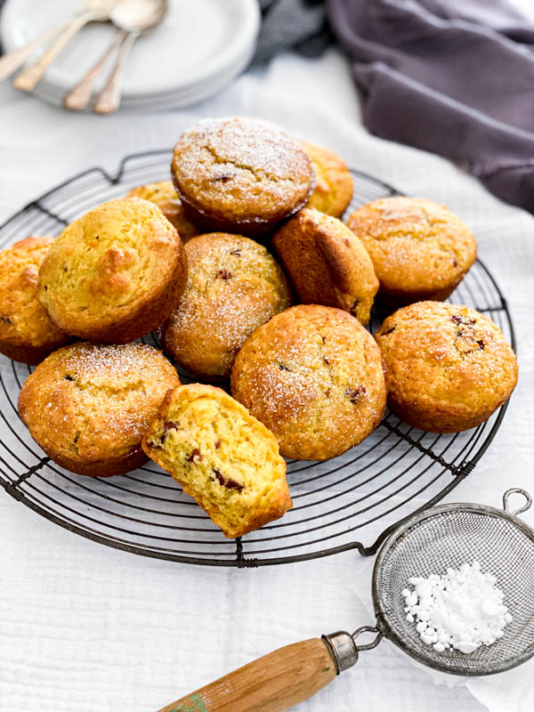 A round wire resting rack has a mound of freshly baked muffins on it. In the background are plates and cutlery. In the foreground is a small sieve with icing sugar in it.