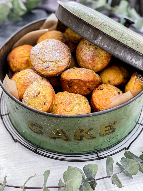 An old vintage mint green cake tin lined with baking paper is filled with muffins and the lid is propped on one side.