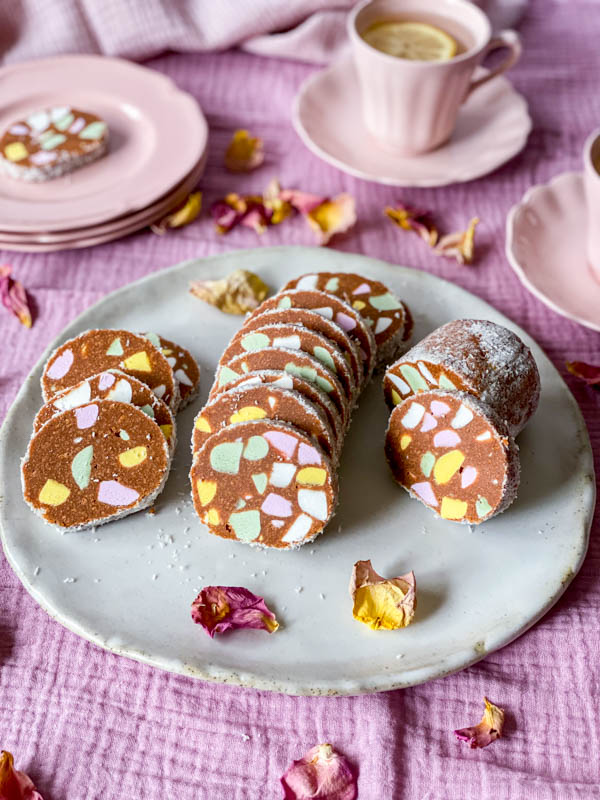 The Lolly Cake slices are on a plate with plates and tea cups int the background. They are set on a pink tablecloth.