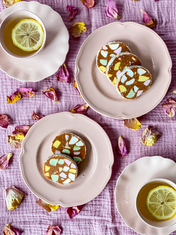 Looking down onto two pink serving plates and two cups and saucers. On the plates there are slices of Lolly Cake. They are set on a pink tablecloth with rose petals scattered around them.