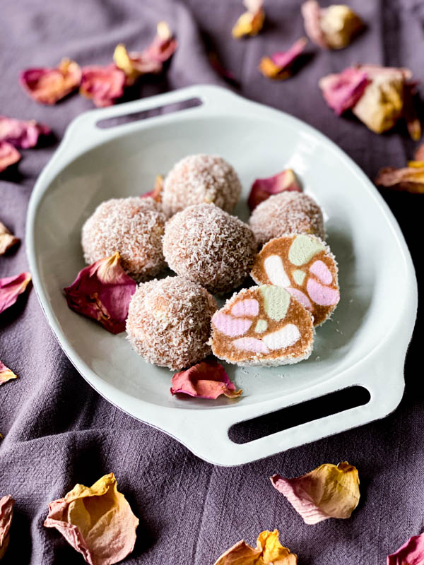 The Lolly Cake mixture is rolled into balls that have been rolled in coconut. The balls are on a plate with one of them sliced open to show the colourful inside. This shows another way to shape the Lolly Cake.