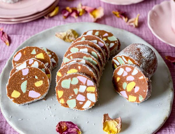 A plate of Lolly Cake slices sitting on a pink tablecloth with pink plates and cups in the background and rose petals scattered around the plates.