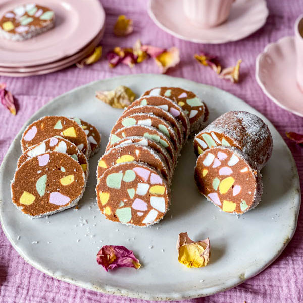 A plate of Lolly Cake slices sitting on a pink tablecloth with pink plates and cups in the background and rose petals scattered around the plates.