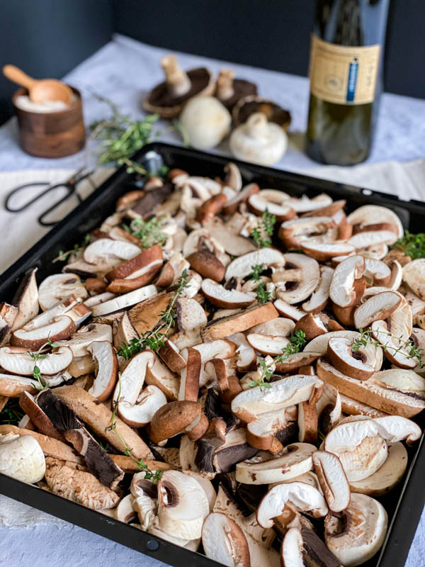 Raw sliced mushrooms are laid out on an oven tray with thyme sprigs sprinkled over. There is a bottle of olive oil in the background along with musrhrooms, herb scissors and a pinch pot of sea salt.