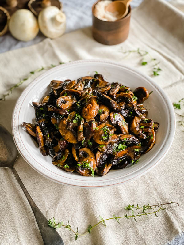 The roasted mushrooms are in a serving bowl sprinkled with parsley. Thyme sprigs are scattered around the bowl and there are whole mushrooms and a pinch bowl of sea salt in the background.