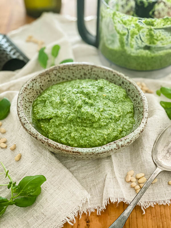 The pesto is in a small ceramic bowl with the mini food processor bowl in the background. Pine-nuts and basil leaves are scattered around the bowl.