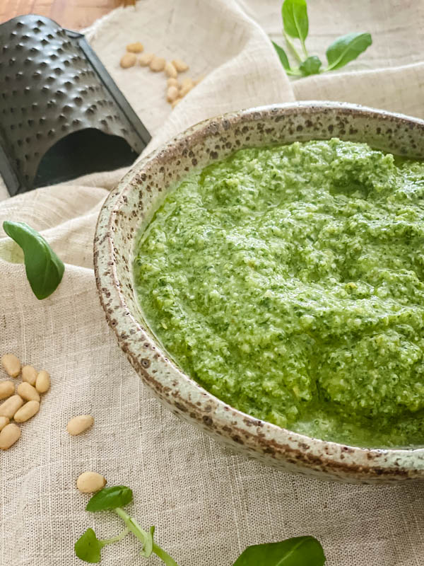 Another side close up of the bowl of Basil Pesto. A mini grater is in the background with pine-nuts and basil leaves scattered around the bowl.