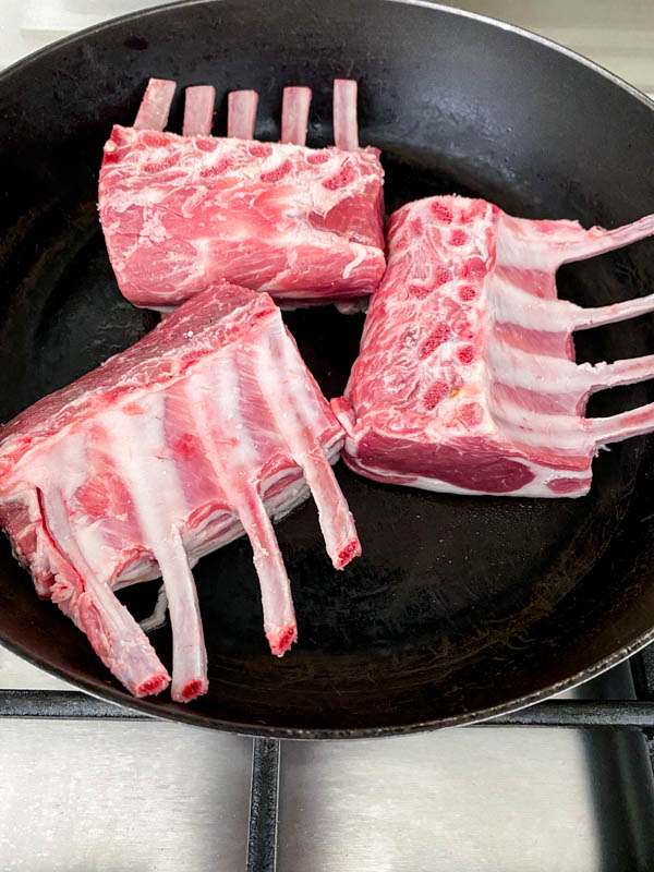 The lamb racks are browning in a frying pan, fat side down.