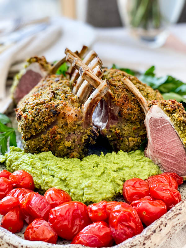 The cooked Herb Crusted Lamb Racks are on a serving platter with one of the racks cut into. They are surrounded by pea puree and blistered baby tomatoes.