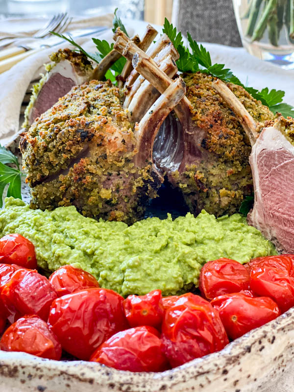 A close up of the Herb Crusted Lamb Racks on a serving platter with pea puree and blistered baby tomatoes.