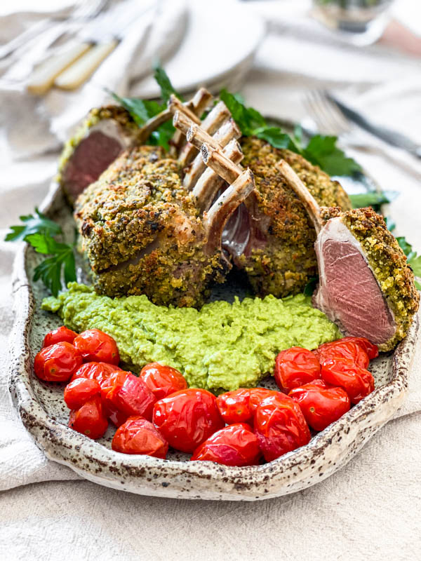 The platter of Herb Crusted Lamb Racks, pea puree and blistered tomatoes are on a table with cutlery and plates in the background.