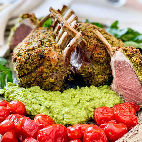 Close up of the cooked lamb racks with pea puree and blistered tomatoes. One of the racks has been cut into showing the beautiful pink lamb meat inside.