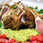 The Herb Crusted Lamb Racks are on a platter ready to be served. There is pea puree and blistered baby tomatoes on the platter as well.