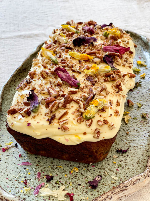 The Banana Cake is on a plate and topped with Cream Cheese Frosting with walnuts and dried flowers on top.