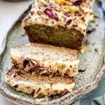 The Banana Cake is on a serving plate and has been frosted with Cream Cheese icing and decorated with walnuts and dried flowers.