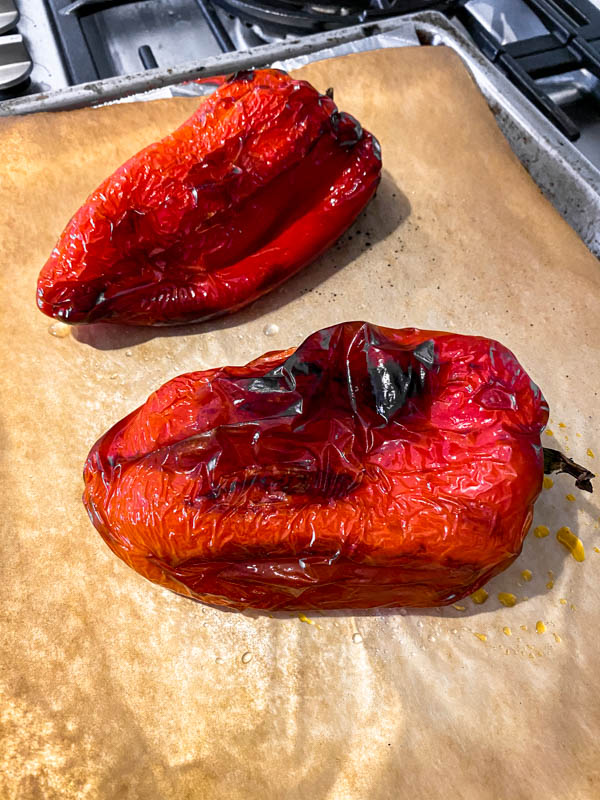2 red capsicums that have been roasted are on an oven tray. Their skin is blackened and they are ready to be peeled.