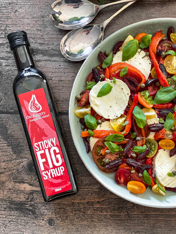 Looking down onto the table with the Caprese Salad, salad servers and a bottle of Sticky Fig Syrup.