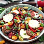 The Caprese Salad is on a wooden table with whole tomatoes and basil leaves around it.