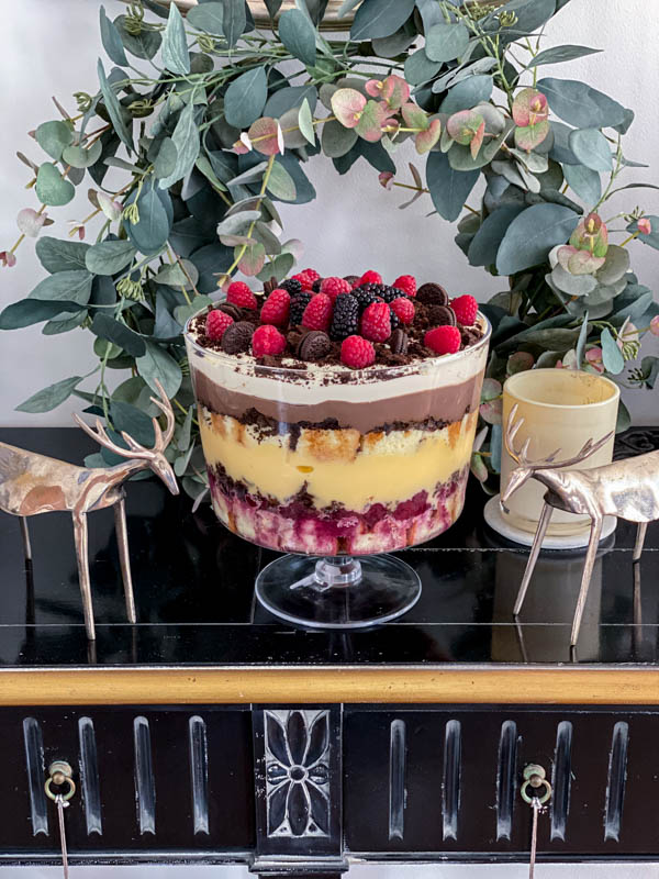The trifle is on a black wooden sideboard with a wreath behind it and Christmas decorations around it.